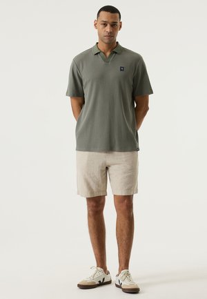 Korte mouwen, olijfgroene poloshirt van textuurstof, met een V-hals en een klein logopatch; gecombineerd met lichtbeige shorts.