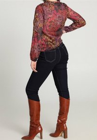 Blusa floreale nei toni del rosso e viola, con maniche lunghe, polsini elastici e colletto arricciato. Abbinata a jeans scuri e stivali marroni al ginocchio.