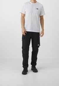 Camiseta blanca de algodón con un pequeño logo negro en el pecho, combinada con pantalones cargo negros que tienen bolsillos laterales y detalles con cremallera.