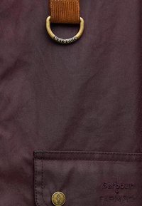 Μπορντό ύφασμα με υφασμάτινη επιφάνεια, διαθέτει καφέ κορδόνι και έναν στρογγυλό χάλκινο δακτύλιο με ανάγλυφο το "Barbour". Περιλαμβάνει λεπτομέρεια κουμπιού.