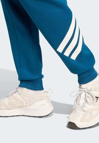 Teal sweatpants feature white stripes on the left leg, paired with white athletic shoes showcasing mesh and smooth textures.