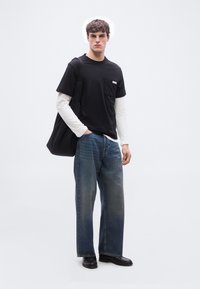 Νέος άνδρας που φοράει στρωμένα μαύρα και λευκά μακρυμάνικα πουκάμισα, φαρδιά μπλε τζιν, μαύρα παπούτσια και κρατάει μια μεγάλη μαύρη τσάντα στον ώμο.