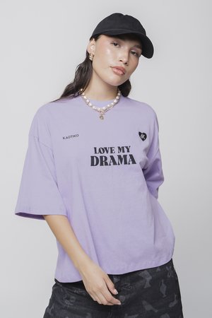 Purple oversized t-shirt with "LOVE MY DRAMA" text in black, short sleeves, and a heart logo. Worn with a pearl necklace and black cap.