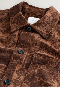 Brown patterned shirt with a collar, featuring intricate dark brown designs, two black buttons, and a label with "NEXT" on the inner neck.