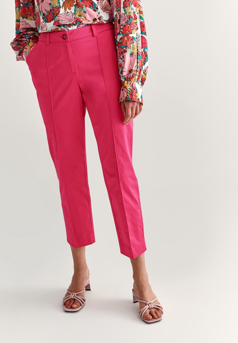 Pantalons rose coupés avec une texture lisse, dotés d'une fermeture à bouton, de poches latérales et de plis marqués le long des jambes avant.