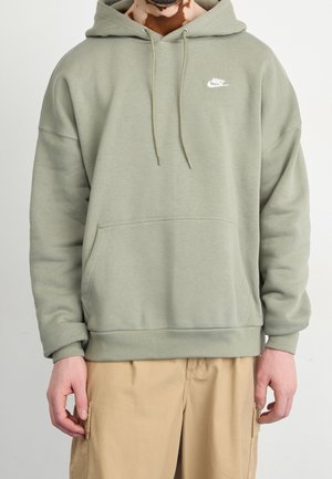 Mężczyzna w jasnozielonej bluzie Nike z kapturem i przednią kieszenią oraz beżowych spodniach, stojący na tle jednolitego jasnego tła.