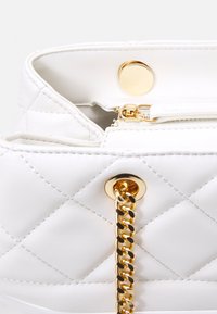 Sac à main blanc matelassé avec quincaillerie dorée, doté d'une fermeture à bouton sur le dessus, d'une poche zippée et d'une bandoulière en chaîne dorée.