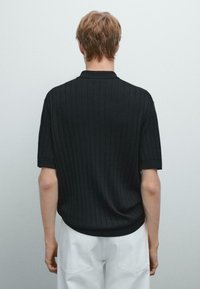 Polo shirt nero a coste con maniche corte e una sottile texture, dotato di colletto e orlo dritto. Indossato con pantaloni bianchi.