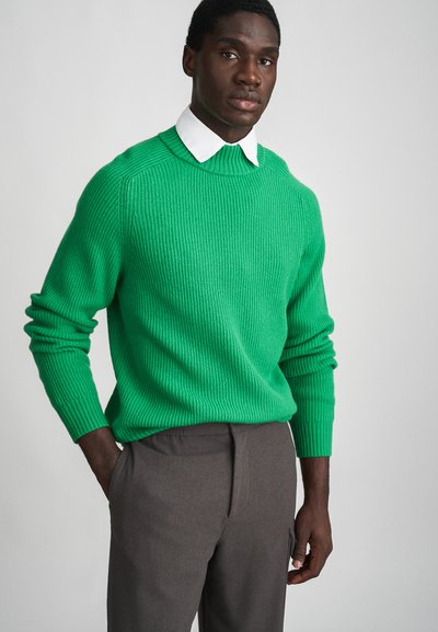 Maglione verde a coste con collo rotondo, maniche lunghe e finitura testurizzata, abbinato a pantaloni grigi.