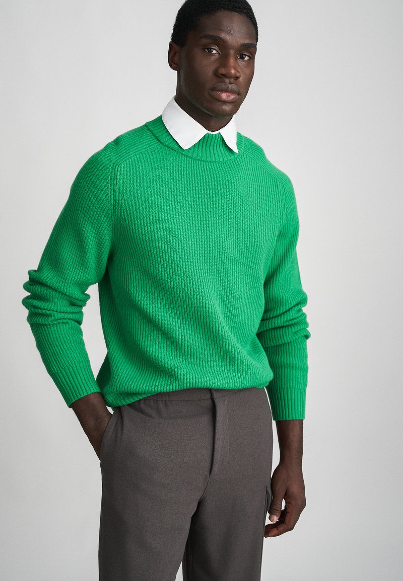 Pull en maille côtelée vert avec un col rond, des manches longues et une finition texturée, associé à un pantalon gris.
