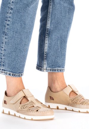 Des chaussures en cuir perforé beige présentent une sangle Velcro, une semelle blanche à rainures et des accents découpés, associées à un jean en denim bleu cropped.