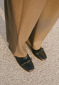 Zapatos cuadrados de cuero negro con acento de hebilla, combinados con pantalones sueltos de textura marrón claro, vistos desde arriba sobre una superficie de grava.