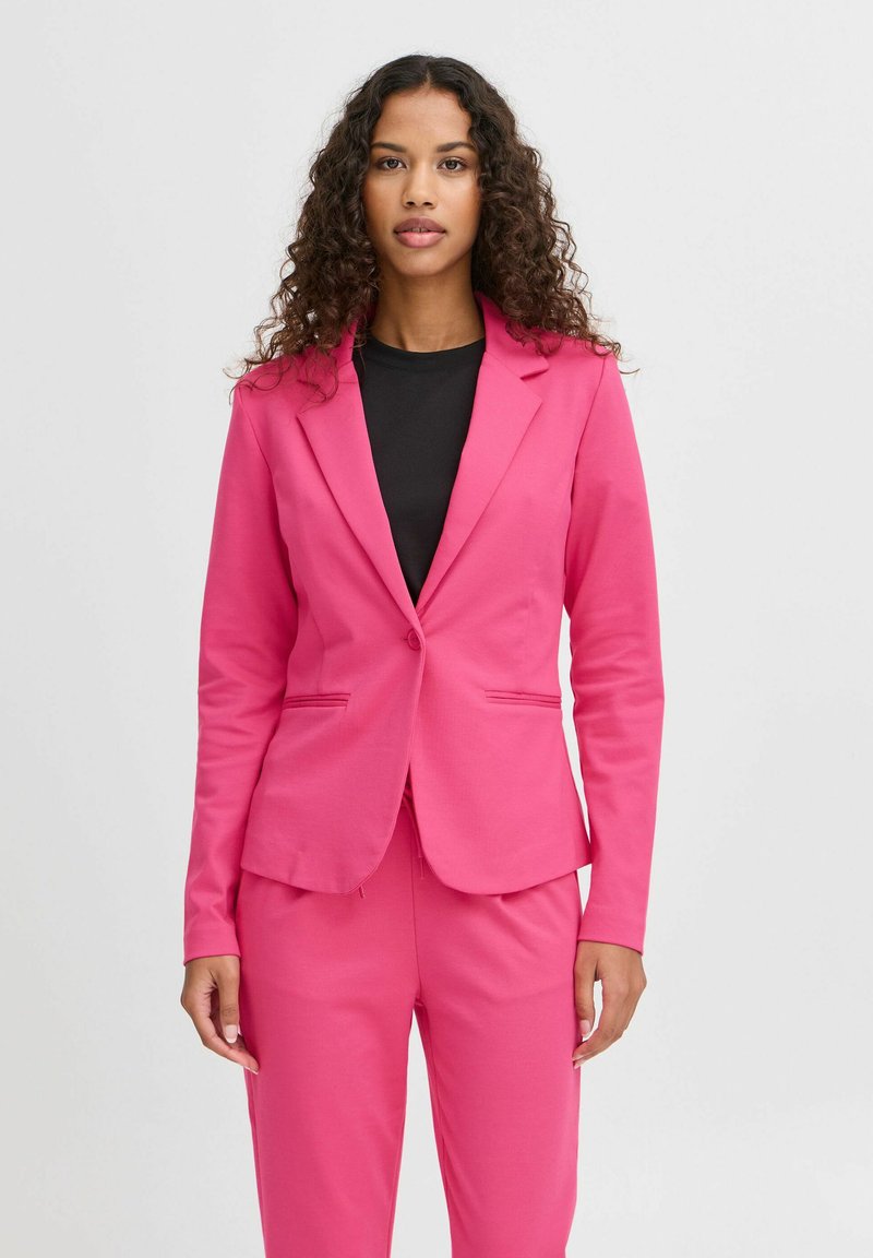 Femme aux cheveux bouclés portant un blazer rose vif ajusté et un pantalon assorti sur un haut noir, debout devant un fond blanc.