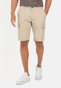 Beige cargoshorts met zijzakken, een platte voorkant en een omslagzoom, gecombineerd met witte sneakers. De stof lijkt licht en casual.