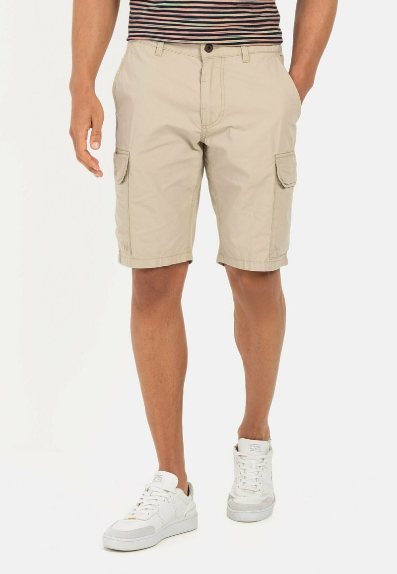 Beige cargoshorts met zijzakken, een platte voorkant en een omslagzoom, gecombineerd met witte sneakers. De stof lijkt licht en casual.