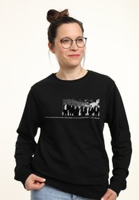 Eine lächelnde Frau trägt einen schwarzen Sweatshirt mit einem Vintage-Foto von Männern in Anzügen und einem Zitat eines beliebten Filmzeichentrickcharakters.