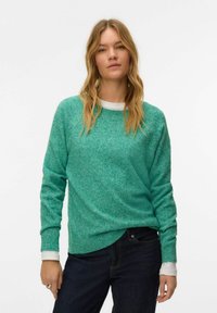 Femme aux longs cheveux blonds portant un pull vert sur un sous-pull blanc à manches longues et un jean bleu foncé, debout devant un fond blanc uni.