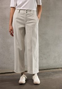 Persoon in hoogstaande beige corduroy wijde broek, witte sneakers en een ingestopt kort mouw wit shirt, staand tegen een betonnen muur.