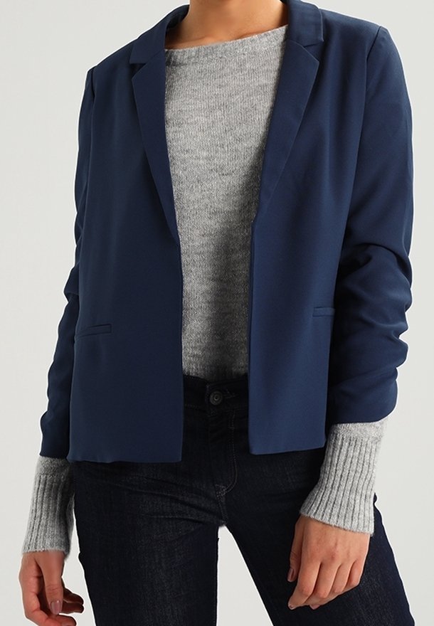 Blazer azul marino con un diseño ajustado, tela suave, solapas con muesca y dos bolsillos frontales, llevado sobre un suéter de punto gris.