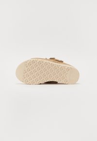 Beige semskede sko med en teksturert gummisåle som har et sirkulært mønster og justerbar stroppdetalj på siden.
