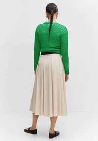 Grüner gestrickter Pullover mit strukturiertem Muster, schwarzem Bund und einem beigen, plissierten Rock. Schwarze Ballerinas runden das Outfit ab.