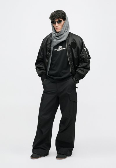 Μαύρο μπουφάν bomber πάνω από μαύρο μπλουζάκι με γραφικά, συνδυασμένο με φαρδιά μαύρα cargo παντελόνια και γκρίζο κασκόλ. Τα αξεσουάρ περιλαμβάνουν σκούρα γυαλιά ηλίου και γάντια.