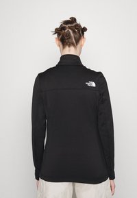 Czarna polarowa kurtka z wysokim kołnierzem, z białym logo na górnej części pleców. Gładka tekstura i dopasowany krój.