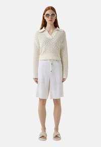 Gebreide crèmekleurige trui met een V-hals en lange mouwen, gecombineerd met witte linnen shorts met twee voorzakken en zilveren hardware. Beigekleurige slipper sandalen.