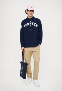 Ναυτικό πουλόβερ με άσπρο κείμενο "YANKEES", κολάρο με κουμπιά; μπεζ cargo παντελόνια; ροζ καπέλο; μπλε τσάντα ώμου; άσπρα αθλητικά παπούτσια.