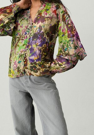 Person iført en gennemsigtig langærmet bluse med blomsterprint og flæsemanchetter samt lysegrå jeans med høj talje mod en ensfarvet baggrund.