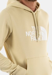 Sudadera beige claro hecha de tejido suave. Cuenta con un bolsillo frontal amplio, capucha con cordones y texto de logo blanco con un diseño curvado.
