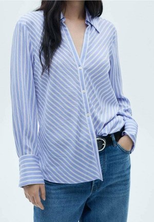 Persona que lleva una camisa de botones a rayas blancas y azul claro metida en vaqueros azules con un cinturón negro, mano en el bolsillo.