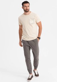 Camiseta de manga corta crema con un bolsillo de camuflaje, combinada con pantalones grises ajustados y zapatillas oscuras con cordones blancos. Atuendo casual.