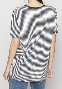 Žena v černobílém horizontálně pruhovaném tričku s krátkým rukávem a modrých džínách, viděná zezadu na jednobarevném pozadí.