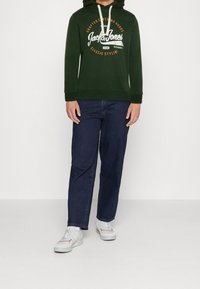 Felpa verde con stampa del logo, cappuccio con lacci, maniche lunghe, abbinata a jeans blu scuro e sneaker bianche. Design semplice e casual.
