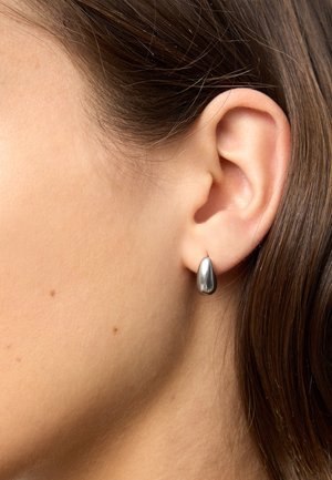Lewe ucho kobiety z gładką srebrną kolczykiem w kształcie kropli łzy oraz brązowe włosy wsunięte za ucho, skóra z widocznymi małymi piegami.