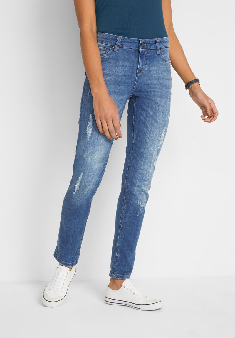 Personne portant un jean bleu déchiré, des baskets blanches et un haut sans manches bleu foncé, debout devant un fond clair uni.