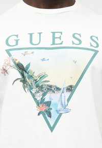 Άσπρο πουκάμισο με τριγωνικό σχέδιο που απεικονίζει τροπικά πουλιά, λουλούδια, ιστιοπλοϊκά και αεροπλάνα, με το κείμενο "GUESS" πάνω από το τρίγωνο.