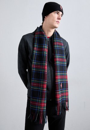 Młody mężczyzna w czarnej czapce typu beanie, ciemnej kurtce zapinanej na zamek oraz długim, kratkowanym szaliku w kolorach czerwonym, zielonym, niebieskim i czarnym, stojący z rękami za plecami.