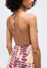 Γυναίκα με σγουρά μαλλιά, ντυμένη με ένα φόρεμα halter tie-dye δίχως πλάτη σε κίτρινους και μαρον αποχρώσεις, εμφανιζόμενη από πίσω με φόντο ένα απλό χρώμα.