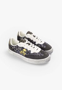 Par de zapatillas de charol en blanco y negro con suelas texturizadas y logo de calavera y tibias en amarillo en los laterales con patrón de camuflaje.
