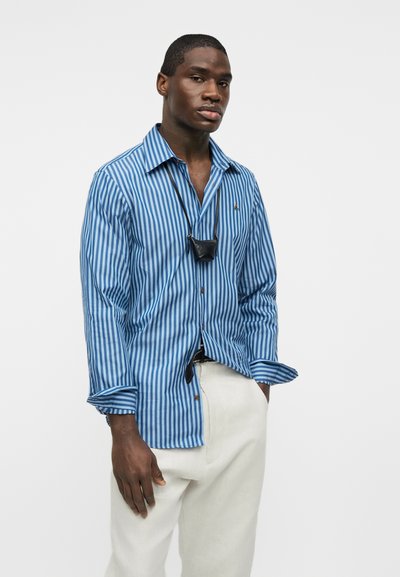 Άνδρας που φοράει μπλε και λευκό πουκάμισο με κάθετες ρίγες, λευκό παντελόνι και μικρό μαύρο περιδέραιο-τσάντα, στέκεται μπροστά σε απλό φωτεινό φόντο.