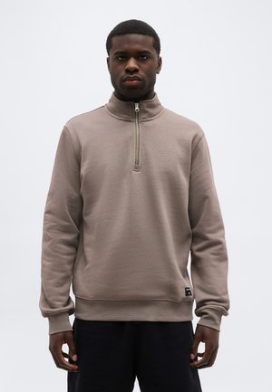 Mann trägt ein taupefarbenes Sweatshirt mit Viertelreißverschluss und dunkle Hose, steht vor einem schlichten hellen Hintergrund und blickt nach vorne.