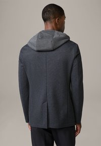 Ciemnoszara kurtka hybrydowa z teksturowaną zewnętrzną warstwą i jasnoszarym kapturem. Posiada wentylację z tyłu oraz dopasowane rękawy.