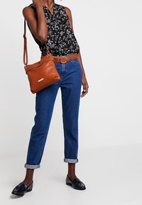 Schwarze Blumenbluse, braune Leder-Schultertasche, blaue Jeans mit umgeschlagenem Saum und schwarze Slipper. Lässiges Outfit mit unterschiedlichen Texturen.