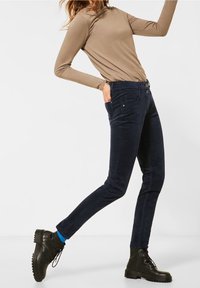 Donkerblauwe ribfluwelen pantalons met een slanke pasvorm, voorzien van twee knopen bij de taille en voorzakken, gecombineerd met zwarte veterschoenen.