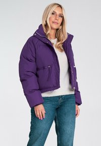 Fioletowa kurtka puchowa z wysokim kołnierzem, zamek błyskawiczny z przodu i zatrzaskami, posiadająca kieszeń na zamek błyskawiczny oraz pikowany wzór.