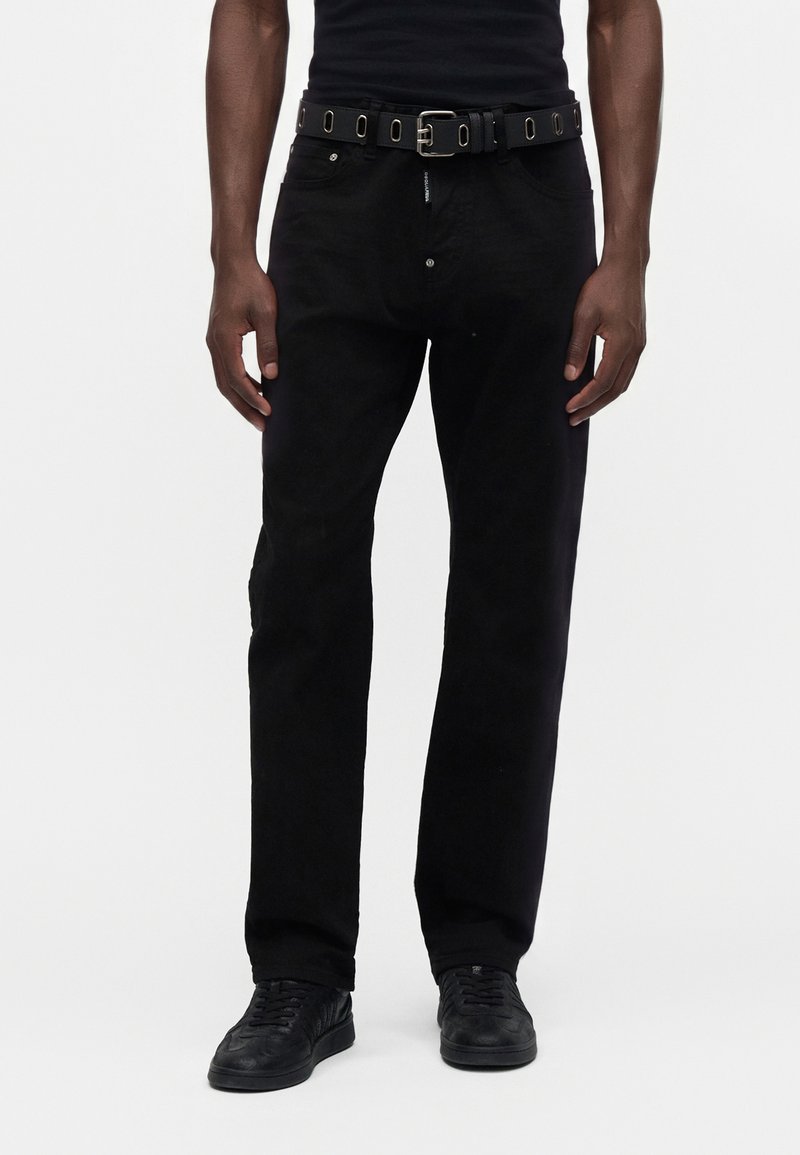 Homme portant un pantalon noir droit, des baskets noires, une ceinture noire avec des œillets métalliques et une chemise noire, debout devant un fond blanc.