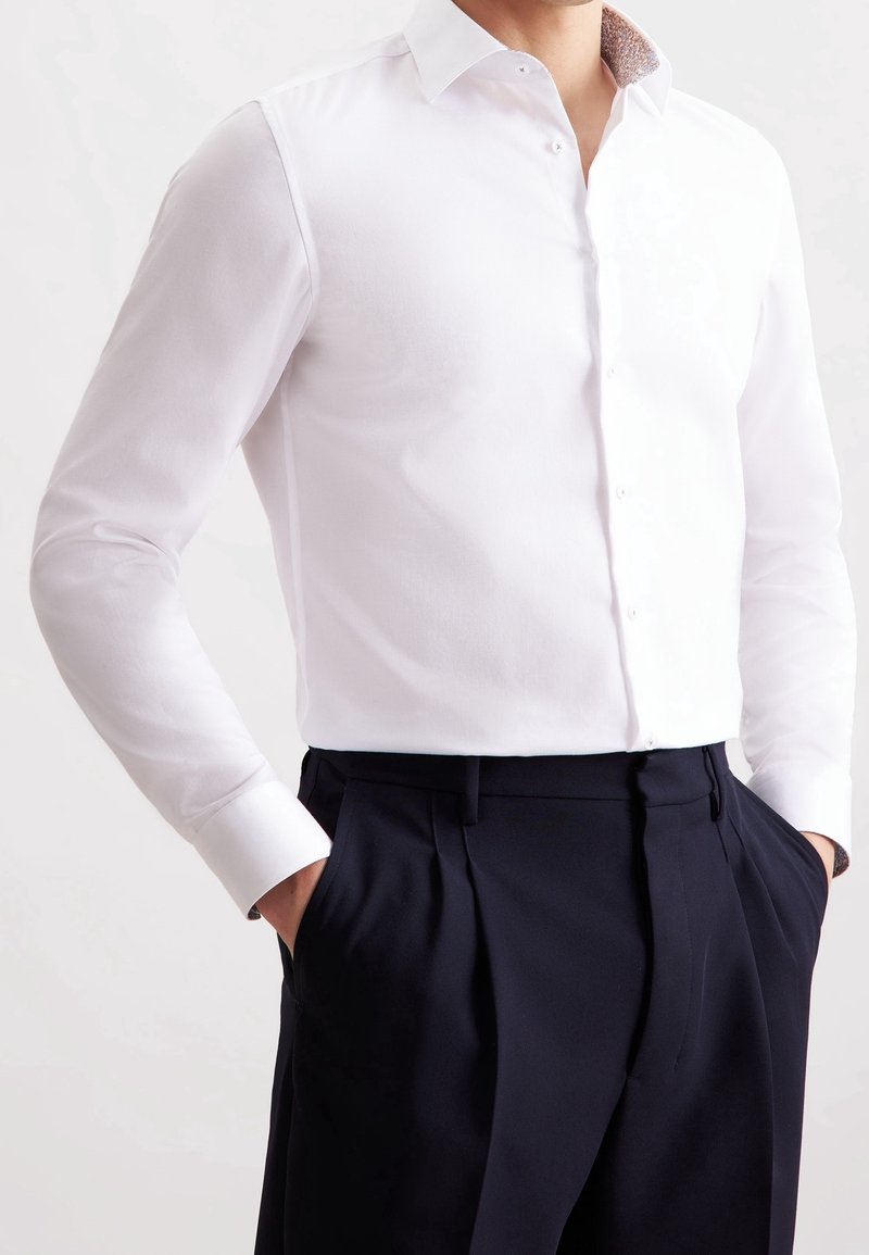 Homme portant une chemise blanche ajustée rentrée dans un pantalon noir plissé taille haute, mains dans les poches.