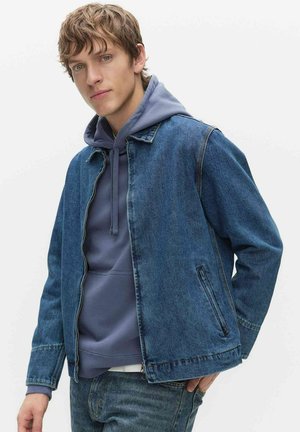 Młody mężczyzna w niebieskiej bluzie z kapturem pod niebieską kurtką dżinsową i dżinsach, stoi z jedną ręką w kieszeni na jednolitym tle.